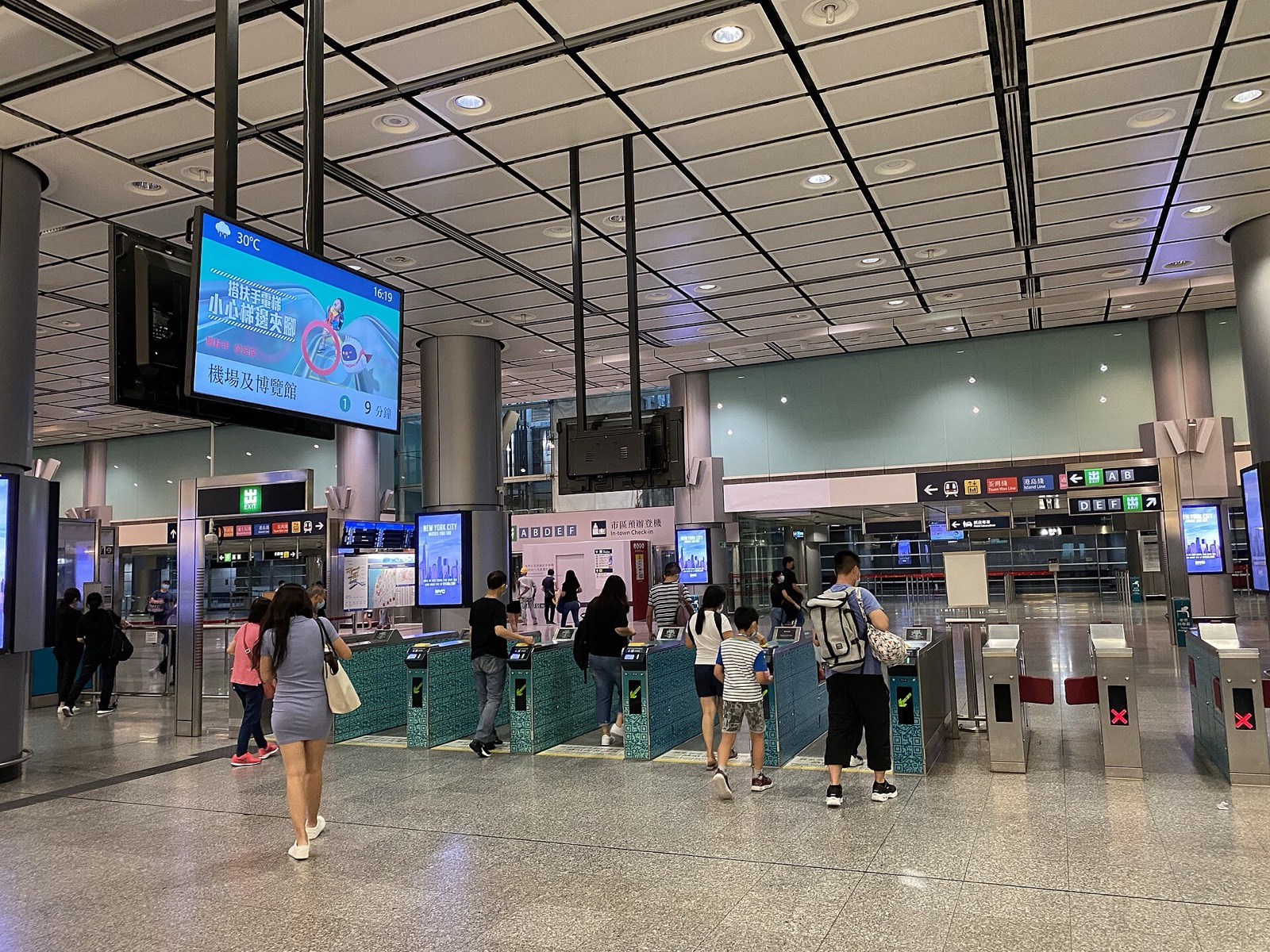 Airport Express fare gates at Hong Kong Station: a row of teal gates with travelers tapping cards to pass through, an overhead 市區預辦登機 In-Town Check-In sign and platform letters A B D E F.