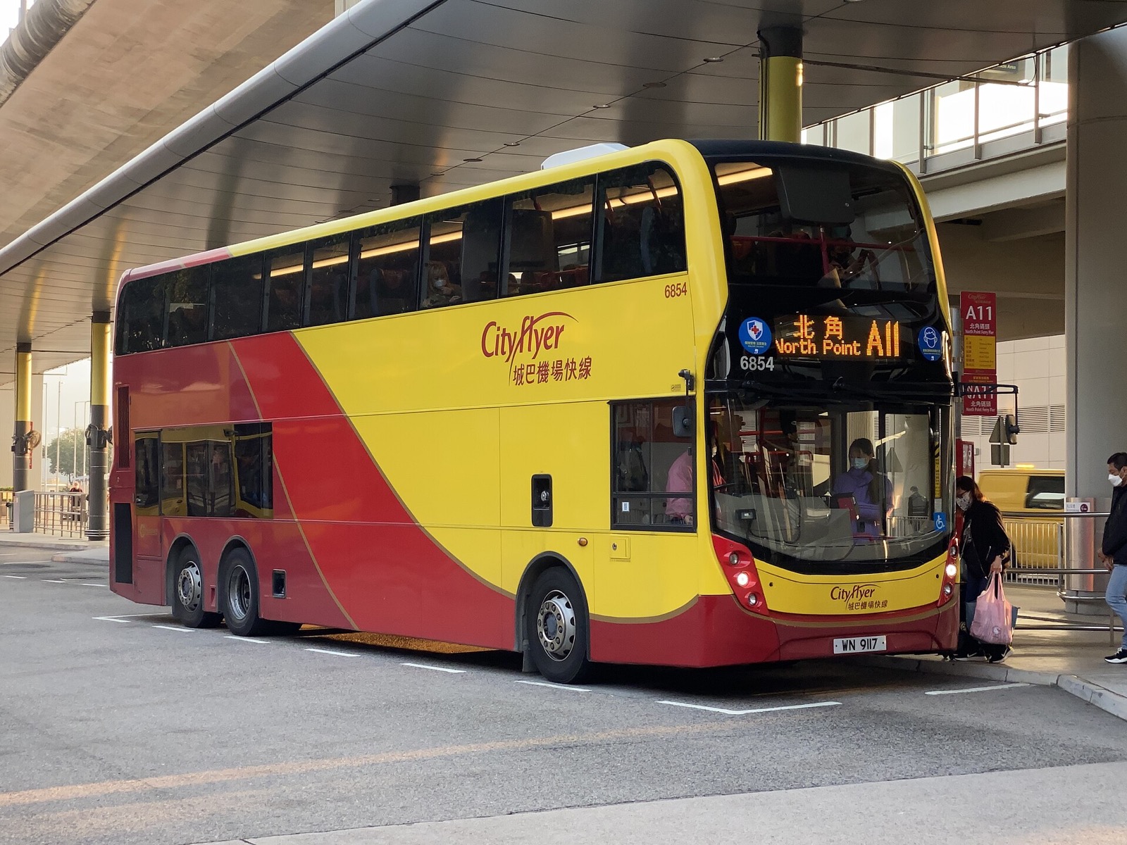 A yellow-and-red Cityflyer double-decker at a covered stand, front display reading North Point 北角 A11, fleet number 6854 and plate WN 9117, a red A11 stop sign behind, and a passenger with a pink bag at the front door.