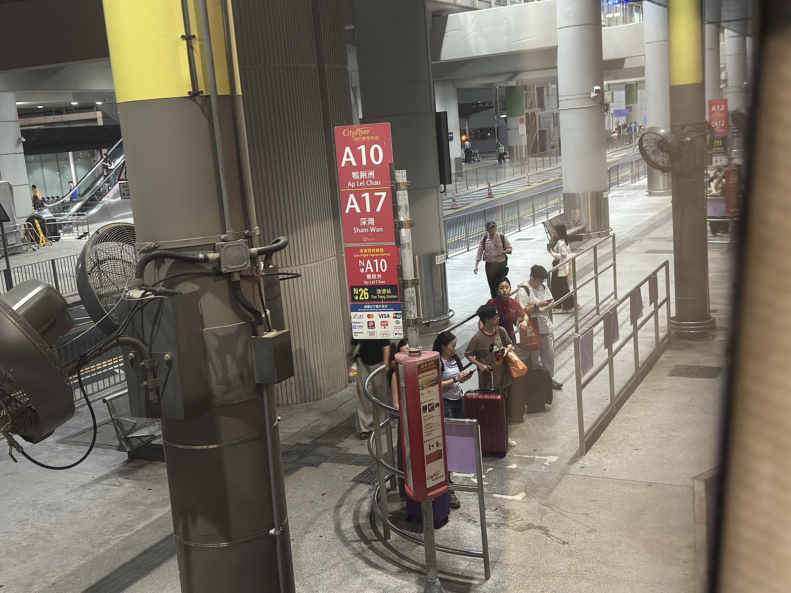 A red CityFlyer route sign at the Ground Transportation Centre bus terminus for A10 Ap Lei Chau / A17 Shum Wan / NA10 / N26 Yam Tong, travelers queuing with luggage, Visa/Mastercard/Apple Pay/Octopus payment icons on the sign.