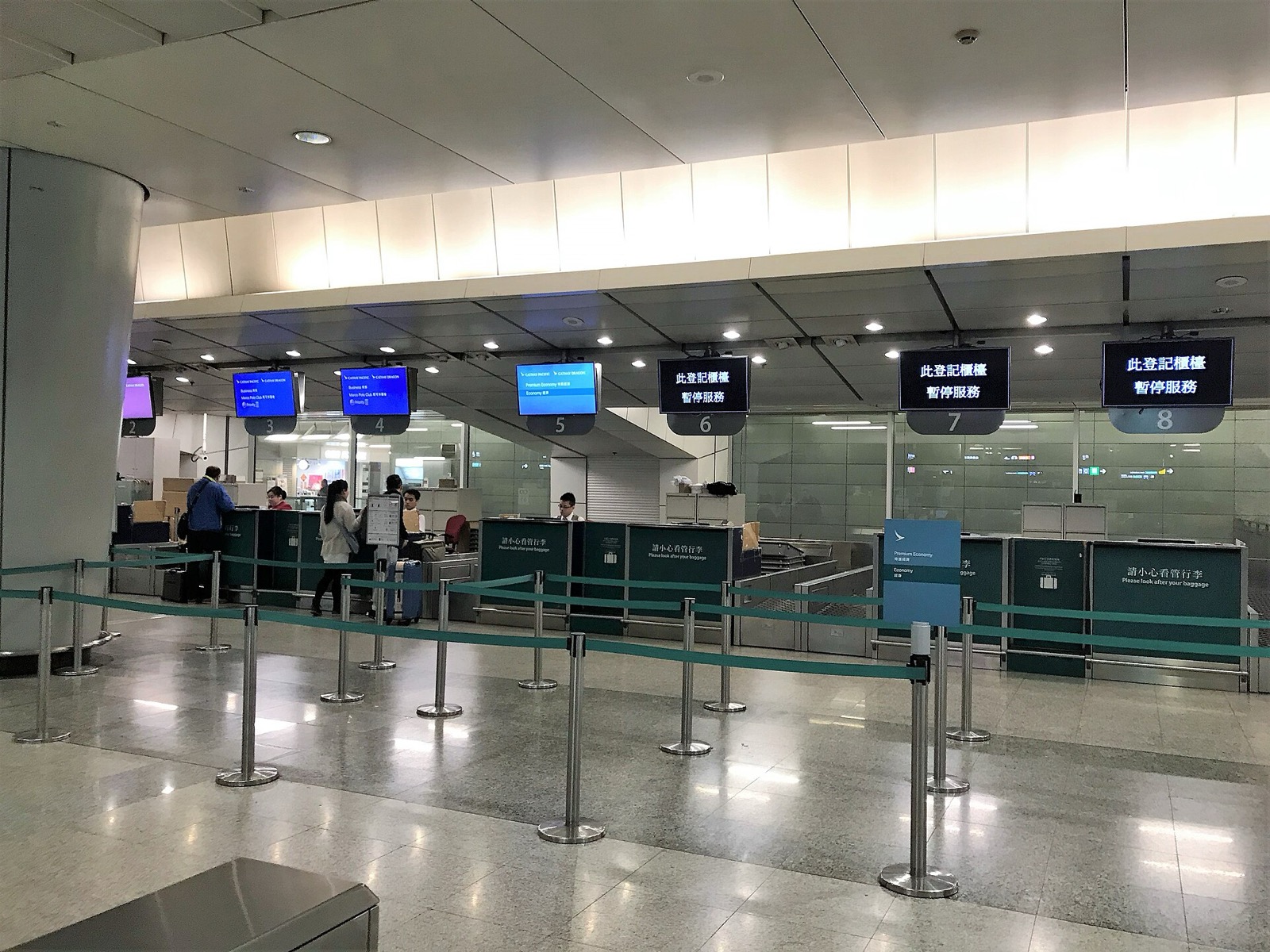 The in-town check-in hall at Kowloon Station: counters 2–5 open with staff and passengers, counters 6–8 marked 'this check-in counter is temporarily suspended', a Cathay Pacific Premium Economy sign to the right.