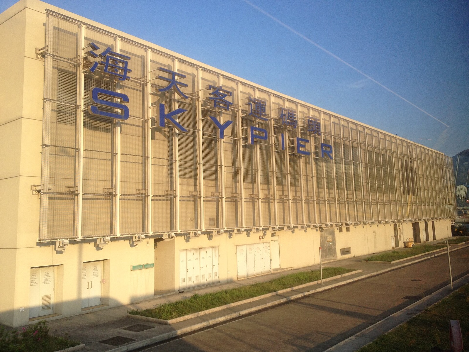 The beige SkyPier building with large blue 海天客運碼頭 and SKYPIER letters on a metal-mesh facade, a paved service road and grass strips in front, no people or boats in frame.