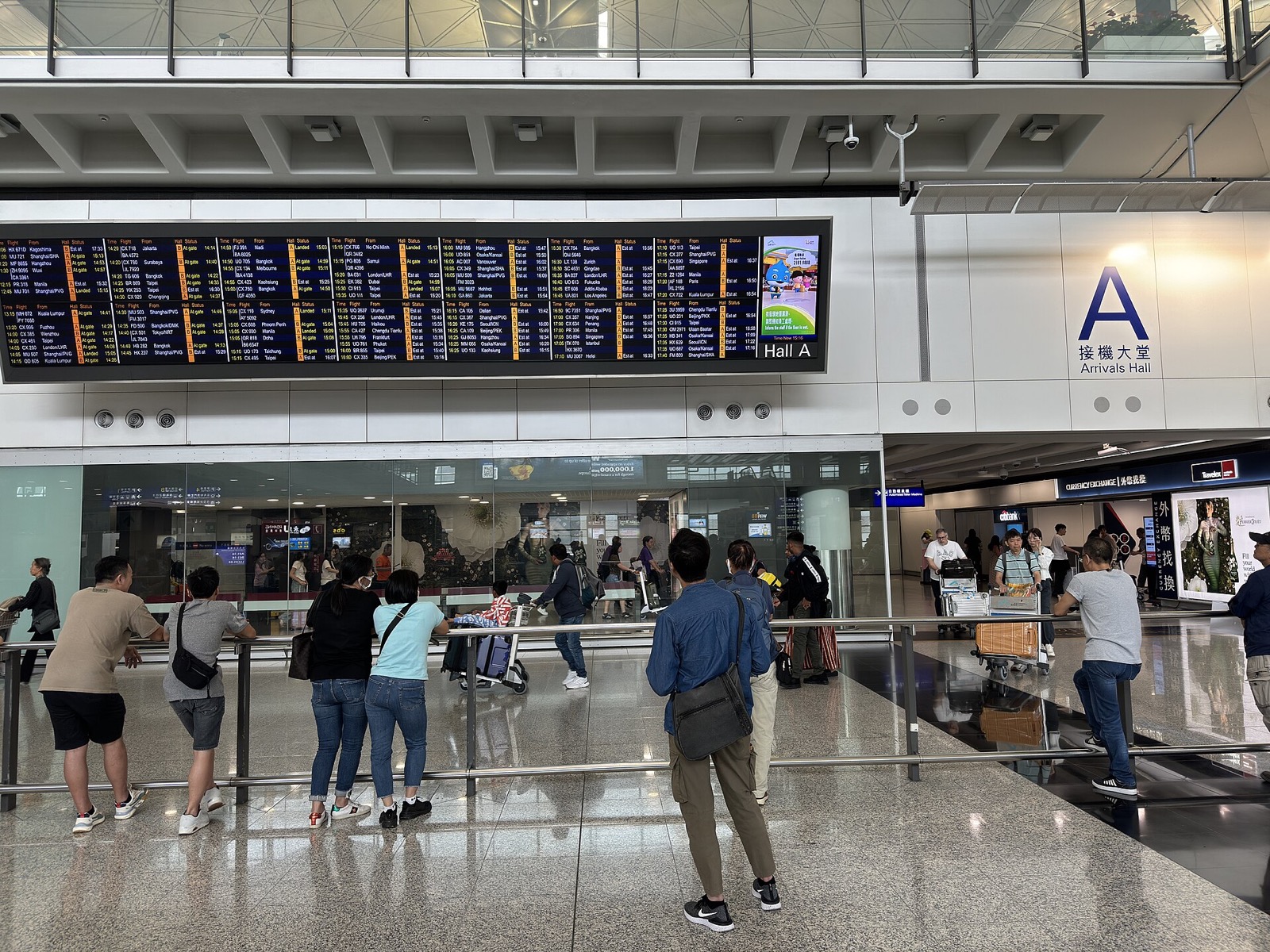 Aéroport international de Hong Kong, Terminal 1, hall des arrivées A : un grand A bleu et 接機大堂 Arrivals Hall sur le mur, un tableau des vols Hall A, des personnes qui attendent à une rampe métallique, des voyageurs avec chariots derrière une vitre, et un bureau de change